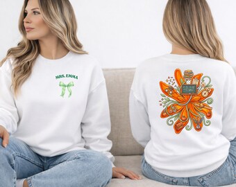 May include: White sweatshirt with a green bow and the text "MRS. EMMA" on the front. The back features a colorful floral design with school-related elements, including books, apples, and a chalkboard. The sweatshirt is a comfortable, casual style.