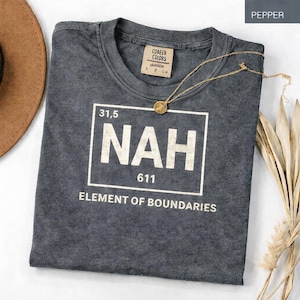 May include: A gray t-shirt with the word "NAH" in a white box, with the text "ELEMENT OF BOUNDARIES" below. A gold necklace with a pendant rests on the shirt. The shirt has a small tag that says "COABER COLORS".