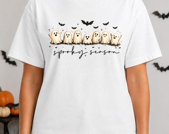 May include: A white short-sleeve t-shirt featuring a graphic print of seven small ghosts in a row with black bats and stars above them. Below the ghosts, the text "spooky season" is written in a black cursive script font across the chest.