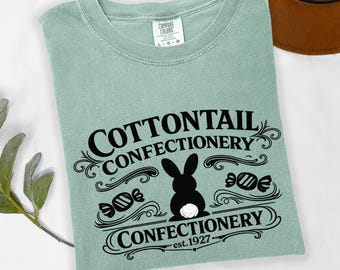 May include: A sage green short-sleeve t-shirt featuring black text that reads COTTONTAIL CONFECTIONERY, a silhouette of a rabbit with a white tail, and the text est. 1927. The shirt has a crew neckline and a Comfort Colors brand label.