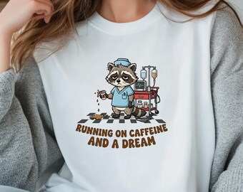 May include: White crew neck t-shirt featuring a cartoon raccoon dressed as a nurse, holding a coffee cup with spilled coffee. The text "RUNNING ON CAFFEINE AND A DREAM" is printed below the raccoon. A medical cart is behind the raccoon.