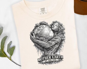May include: Cream-colored t-shirt featuring a detailed black and white illustration of Mother Earth cradling the globe. The design includes the text "MOTHER EARTH WE ARE ONE" in a banner. The shirt is folded.