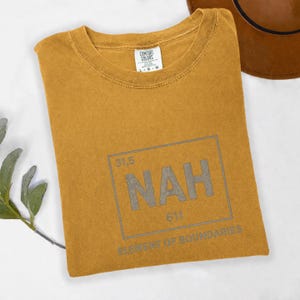May include: Mustard-colored t-shirt with the word "NAH" in a gray, periodic table-style design. The shirt is folded, with the Comfort Colors tag visible. A brown hat is partially visible in the background.