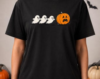 May include: A black short-sleeve t-shirt featuring a graphic print of three small white ghosts chasing a large orange pumpkin with a frightened facial expression. The design is centered on the chest area of the garment, showcasing a playful Halloween theme.