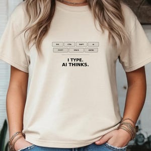 May include: A beige t-shirt with a keyboard layout graphic and the text "I TYPE. AI THINKS." The design includes labels like ESC, CTRL, SHIFT, AI, PROMPT, SPACE, and ENTER, suggesting a tech or computer-related theme.