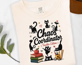 May include: Cream-colored t-shirt with the words "Chaos Coordinator Teacher Edition" in black script, surrounded by cartoon black cats, hearts, stars, books, and an apple. The shirt is made of a soft material.