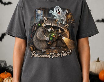 May include: A charcoal gray t-shirt featuring a graphic of two raccoons wearing sunglasses, one holding a ghost-hunting device and the other a flashlight, with a ghost floating above them. The text Paranormal Trash Patrol is printed below.