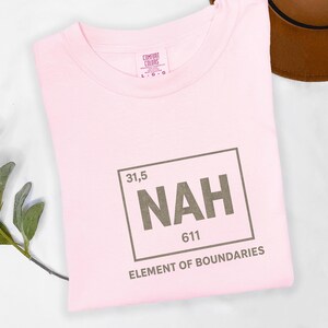 May include: A light pink t-shirt with a crew neck. The shirt features a graphic in a brown rectangle with the word "NAH" in large letters, along with the numbers "31,5" and "611". Below the graphic, the text reads "ELEMENT OF BOUNDARIES".