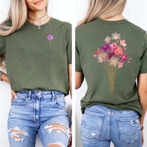 May include: Olive green t-shirt with a small purple flower on the front and a bouquet of pink, purple, and white flowers on the back. The flowers have gold stems. The shirt is paired with blue jeans.