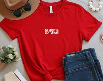 May include: A red t-shirt with the text "YOU DESERVE A GENTLEMAN" in white. The shirt is paired with blue jeans, a brown hat, sunglasses, and white shoes. A white beaded bracelet and a small bouquet of white flowers are also visible.