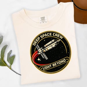 May include: Cream-colored t-shirt featuring a circular graphic with the text "DEEP SPACE CREW" and "LIGHT BEYOND." The graphic depicts a rocket launching against a starry background. The shirt is folded.