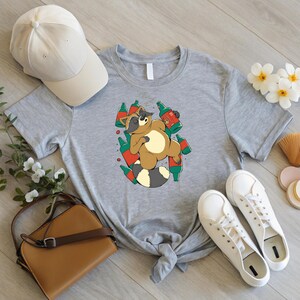 May include: A light gray t-shirt featuring a cartoon raccoon surrounded by beer bottles. The raccoon is brown, tan, and black, with a white belly and a striped tail. The bottles are green and red. A beige baseball cap and white sneakers are also visible.