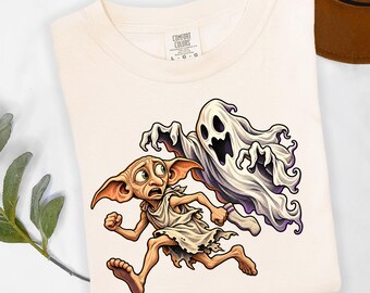 May include: A cream-colored t-shirt featuring a graphic print of a small, large-eared creature wearing a white cloth and running away from a white, translucent ghost. The shirt has a Comfort Colors brand label at the inner neckline.