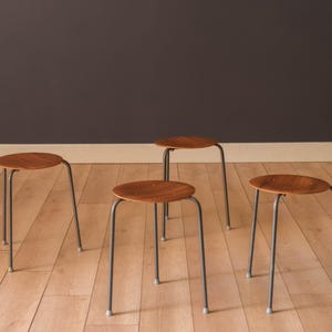 May include: A set of four stools, each featuring a circular wooden seat with a warm brown grain pattern and three slender, dark grey metal legs. The stools are arranged in a group, showcasing their simple, functional design and sturdy construction.