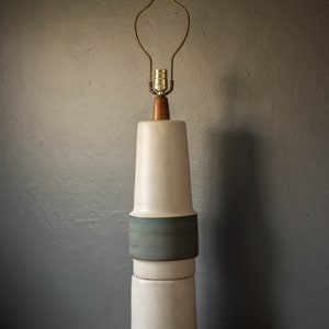 May include: A white ceramic table lamp with a wooden base and a brass wire shade. The lamp has three sections with a green band around each section.