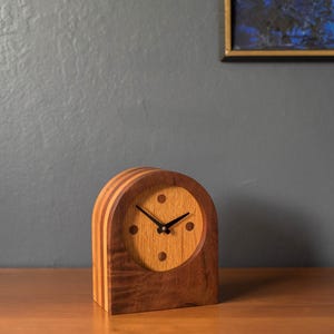 May include: A wooden clock with a curved top and a light brown face. The clock has a dark brown frame and a light brown face with black hands. The clock is on a wooden surface.