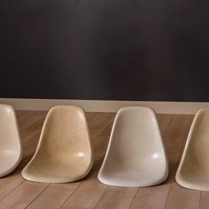 May include: Four chairs in different shades of beige, arranged in a row on a wooden floor. The chairs are made of fiberglass and have a simple, modern design.