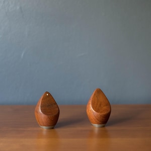 May include: Two wooden salt and pepper shakers with a teardrop design. The shakers are made of a warm-toned wood and have a small, silver-colored base. One shaker has a small white dot on top.