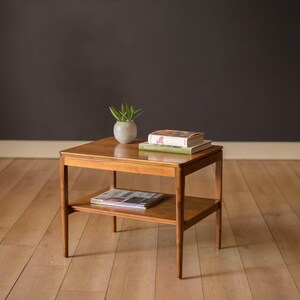 May include: A square, wooden side table with a lower shelf. The table has a glossy finish and holds a small potted plant, two books, and a magazine. The table is approximately 24 inches wide and 24 inches long.