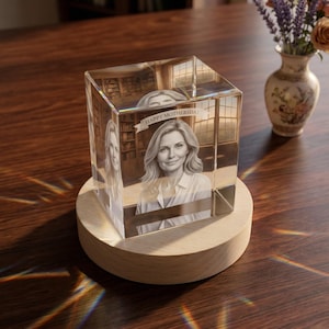 Op de afbeelding: Een helder, rechthoekig kristallen blok met een portret van een vrouw en de tekst "HAPPY MOTHERSDAY". Het kristal staat op een ronde, houten basis. Een vaas met bloemen staat op de achtergrond.