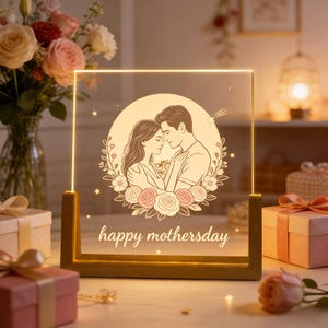 Puede incluir: Panel acrílico cuadrado iluminado con base dorada. Presenta una ilustración de una pareja abrazada dentro de una corona floral, con las palabras "happy mothersday" debajo. El fondo incluye flores y cajas de regalo.