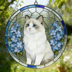 Puede incluir: Una vidriera redonda con un gato Ragdoll de ojos azul brillante y pelaje blanco y gris. El gato está rodeado de flores de hortensia azules. La ventana tiene un borde de vidrio azul y transparente y está suspendida por una cadena.