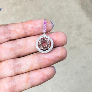 Può includere: Primo piano di un singolo orecchino con una grande gemma rotonda color pesca circondata da un alone di piccole pietre trasparenti. La sezione superiore dell'orecchino ha una fila di piccole pietre rosa. L'orecchino è tenuto in una mano.
