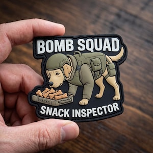 May include: A black rubber morale patch featuring a cartoon yellow dog wearing a tactical olive green vest and helmet. The dog is looking at a tray of bone-shaped treats labeled SNACKS. The text BOMB SQUAD appears above and SNACK INSPECTOR below.