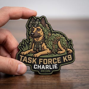 May include: A rubber morale patch featuring a tan and brown dog wearing a green ghillie suit, night vision goggles on its head, and binoculars around its neck. The text reads TASK FORCE K9, CHARLIE, and RECON in bold, white, and tan lettering.