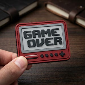 May include: A rectangular patch with a dark red border and a light gray center. The text GAME OVER is embossed in black pixelated font. Below the text, the patch features a horizontal bar, four circular buttons, and a plus sign in relief.