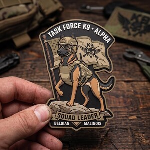 May include: A tactical morale patch featuring a Belgian Malinois dog wearing a helmet and vest, standing on a rock in front of a flag. The text reads TASK FORCE K9 ALPHA, SQUAD LEADER, and BELGIAN MALINOIS. The design includes a skull and crossbones logo.