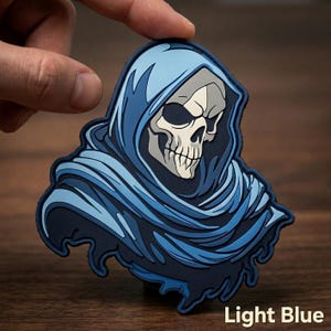 May include: A rubberized patch featuring a skull wearing a hooded cloak in shades of light blue and dark blue. The skull is off-white with dark eye sockets and a detailed jawline. The text Light Blue is printed in the bottom right corner.