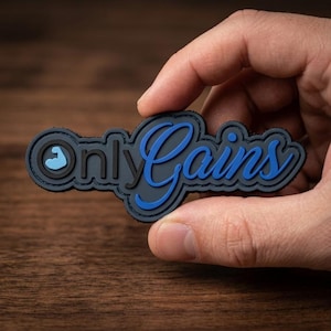 May include: A rectangular, dark gray rubberized patch featuring the text OnlyGains in a raised, bright blue script font. The letter O is replaced by a circular icon containing a light blue flexed bicep silhouette. The patch has a black border.