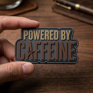 May include: A rectangular PVC morale patch with a dark gray background and a black border. The text POWERED BY is in tan, and the word CAFFEINE is in brown. The patch features a raised, 3D-style lettering design with a clean, bold sans-serif font.