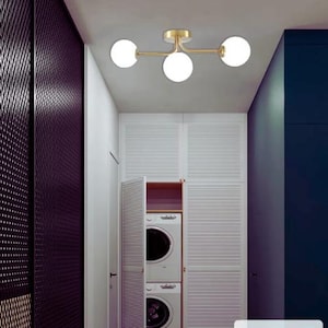May include: A contemporary ceiling light with three spherical white glass shades and a gold-coloured metal frame. The light is mounted on a white ceiling in a room with white and dark blue walls. The room also features a white cabinet and a dark purple wall.
