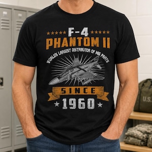 May include: A black short-sleeved t-shirt featuring a graphic of an F-4 Phantom II jet. The text reads F-4 PHANTOM II, WORLD'S LARGEST DISTRIBUTOR OF MIG PARTS, SINCE 1960. The design uses orange and white distressed lettering with star accents.