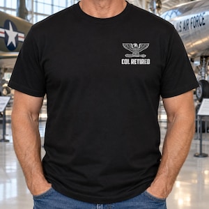 May include: A black short-sleeved t-shirt featuring a white graphic on the upper left chest. The graphic displays an eagle emblem above the text COL RETIRED. The shirt has a crew neckline and is worn with blue denim jeans in a casual style.