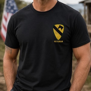 May include: Black crew neck t-shirt with a yellow and black emblem on the left chest. The emblem features a black horse head over a black diagonal stripe on a yellow shield. The word "VETERAN" is printed below the emblem.