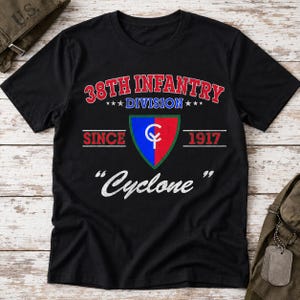Può includere: Una t-shirt nera a maniche corte con il testo 38TH INFANTRY DIVISION SINCE 1917 e la parola Cyclone in bianco. Un emblema centrale a forma di scudo mostra uno sfondo diviso in blu e rosso con un monogramma CY bianco e un bordo verde.