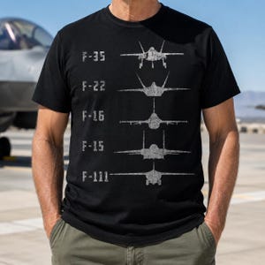 May include: A black short-sleeved t-shirt featuring a white distressed graphic print of five fighter jets arranged vertically. Each jet is labelled with its model name: F-35, F-22, F-16, F-15, and F-111, displayed in a clean, sans-serif font.