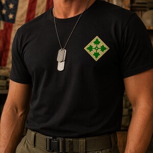 May include: Black t-shirt with a green and tan diamond-shaped emblem on the upper left chest. The emblem features a four-leaf clover design. A silver dog tag necklace hangs from the neck. The background includes an American flag.