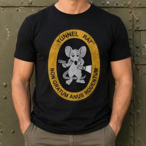 May include: A black short-sleeve t-shirt featuring a central yellow oval emblem with a distressed texture. Inside the emblem is a gray cartoon mouse holding a flashlight and a pistol, with the text TUNNEL RAT NON GRATUM ANUS RODENTUM.