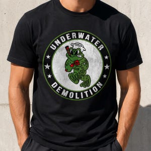 May include: A black short-sleeve t-shirt featuring a circular graphic with a green frog wearing a white sailor hat, holding a cigar and a stick of dynamite. The text "UNDERWATER DEMOLITION" is printed in white around the circular border.