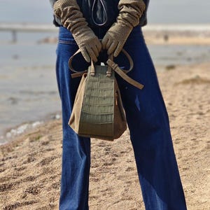 Puede incluir: Un bolso verde oliva con textura de cocodrilo es sostenido por una persona con guantes color beige. El bolso tiene una base de ante marrón y una correa larga. La persona lleva vaqueros azules y zapatos azules.
