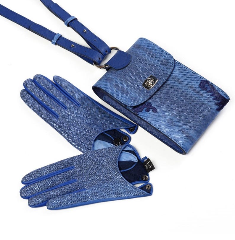 Puede incluir: Un conjunto compuesto por un bolso bandolera azul texturizado con cierre de metal plateado y un par de guantes azules texturizados a juego. El bolso cuenta con una correa larga ajustable y los guantes tienen una peque&ntilde;a etiqueta negra con texto en blanco.