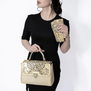 May include: A beige handbag with a gold leopard print flap and handle, paired with matching gloves. The bag has a rectangular shape with a textured body and a metal clasp. The gloves have a similar leopard print design.