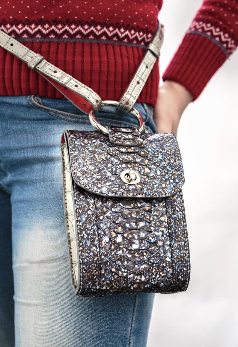 Puede incluir: Un bolso bandolera rectangular vertical con una textura de piel de serpiente iridiscente oscura con motas met&aacute;licas azules y plateadas. El bolso tiene un cierre giratorio de tono plateado y est&aacute; sujeto a una anilla conectada a una correa fina.