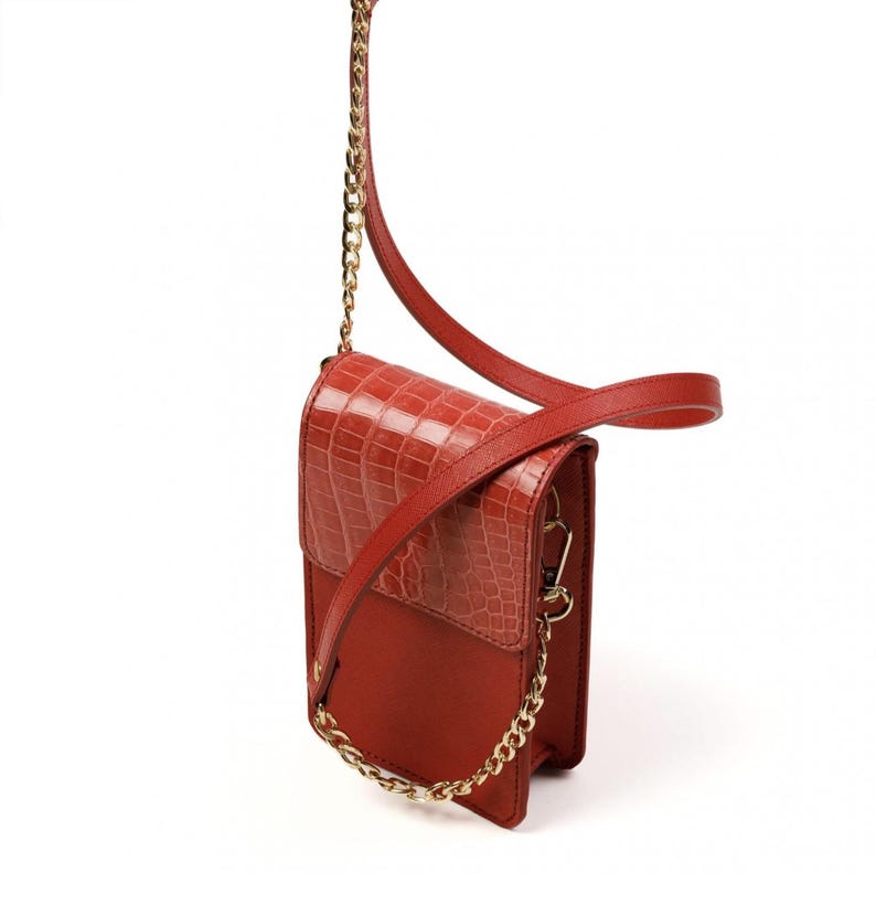Puede incluir: Un bolso bandolera rectangular vertical de color rojo intenso. El bolso presenta una solapa con un patr&oacute;n texturizado de cocodrilo, un cuerpo liso mate, un detalle de cadena de metal dorado en la parte delantera y una correa de hombro roja a juego.