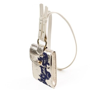 Puede incluir: Un bolso bandolera de color dorado met&aacute;lico con relieve de piel de serpiente y un patr&oacute;n floral bordado en azul oscuro en la solapa frontal. Incluye un cierre de giro dorado, una anilla met&aacute;lica dorada y una correa larga de cuero blanco ajustable.