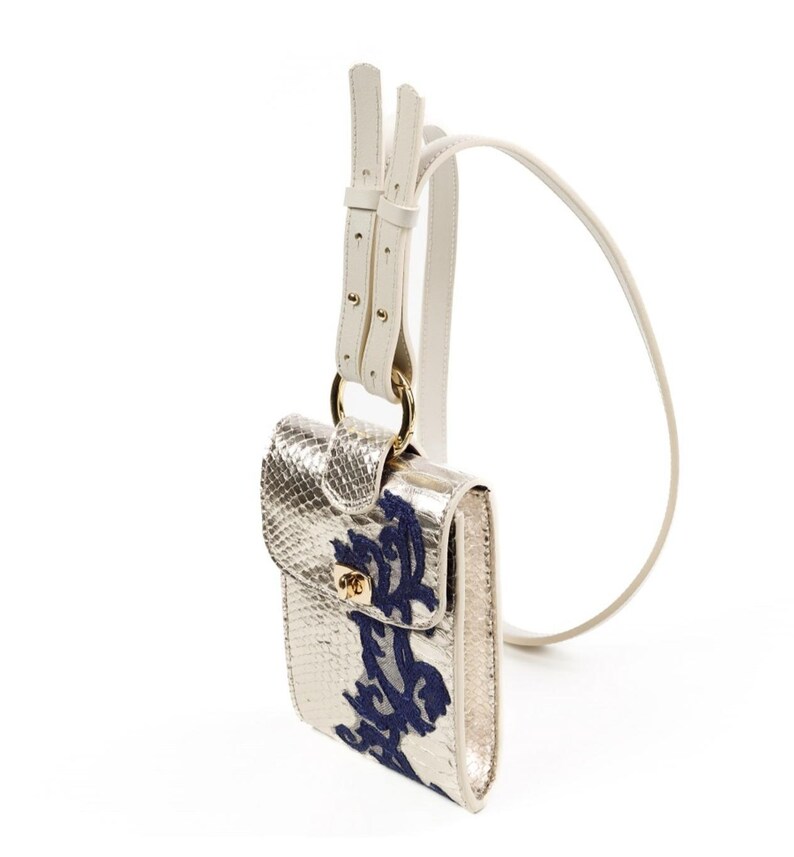 Puede incluir: Un bolso bandolera de color dorado met&aacute;lico con relieve de piel de serpiente y un patr&oacute;n floral bordado en azul oscuro en la solapa frontal. Incluye un cierre de giro dorado, una anilla met&aacute;lica dorada y una correa larga de cuero blanco ajustable.
